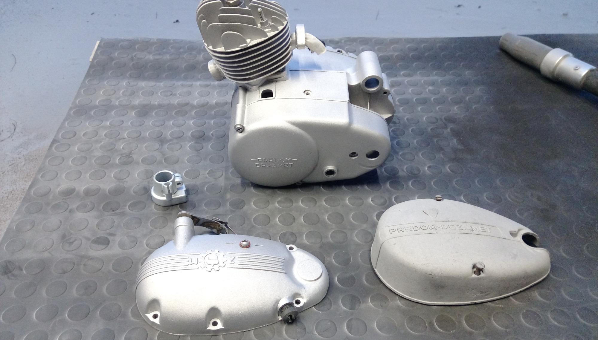 Elementy silnika motocyklowego Predom-Dezamet po renowacji, pokryte srebrzystą farbą, leżące na gumowej macie. Widoczny cylinder, karter i dwie pokrywy.