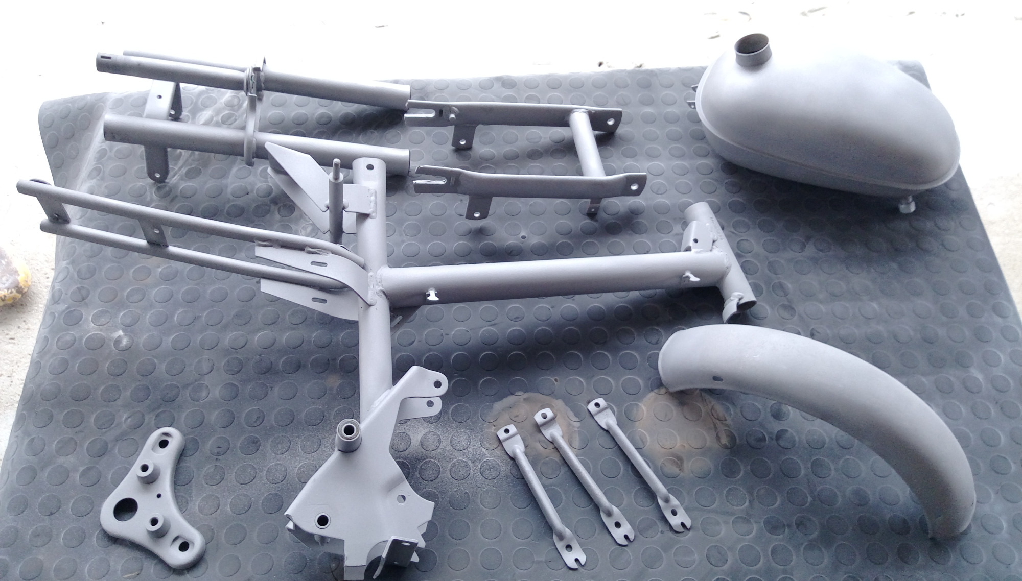 Elementy ramy motocykla i zbiornik paliwa po obróbce, pokryte szarym podkładem, leżące na gumowej macie z okrągłymi wypustkami.