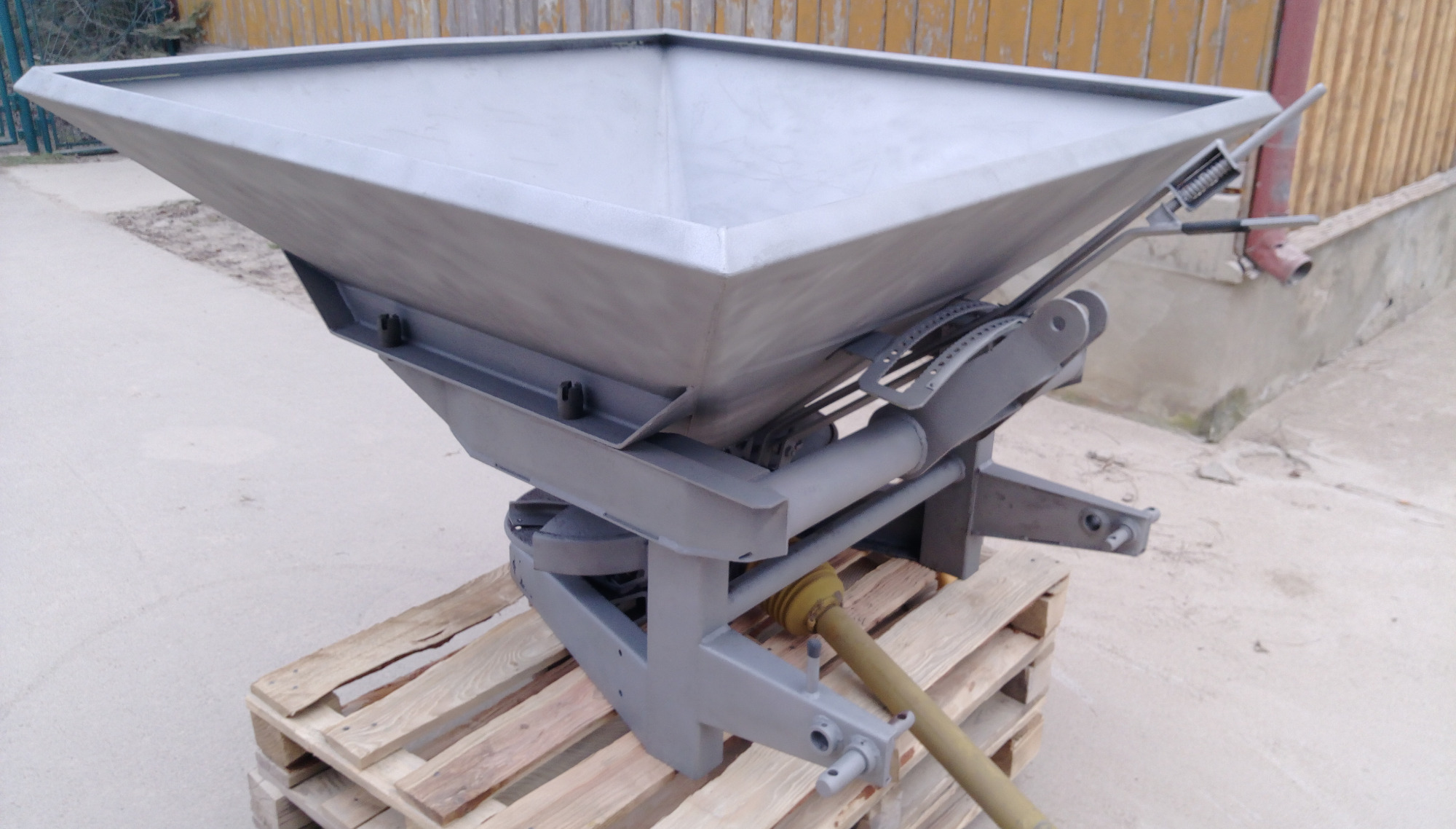 Metalowy rozsiewacz nawozów po renowacji, stojący na palecie, z widocznym mechanizmem regulacji i wałem odbioru mocy.