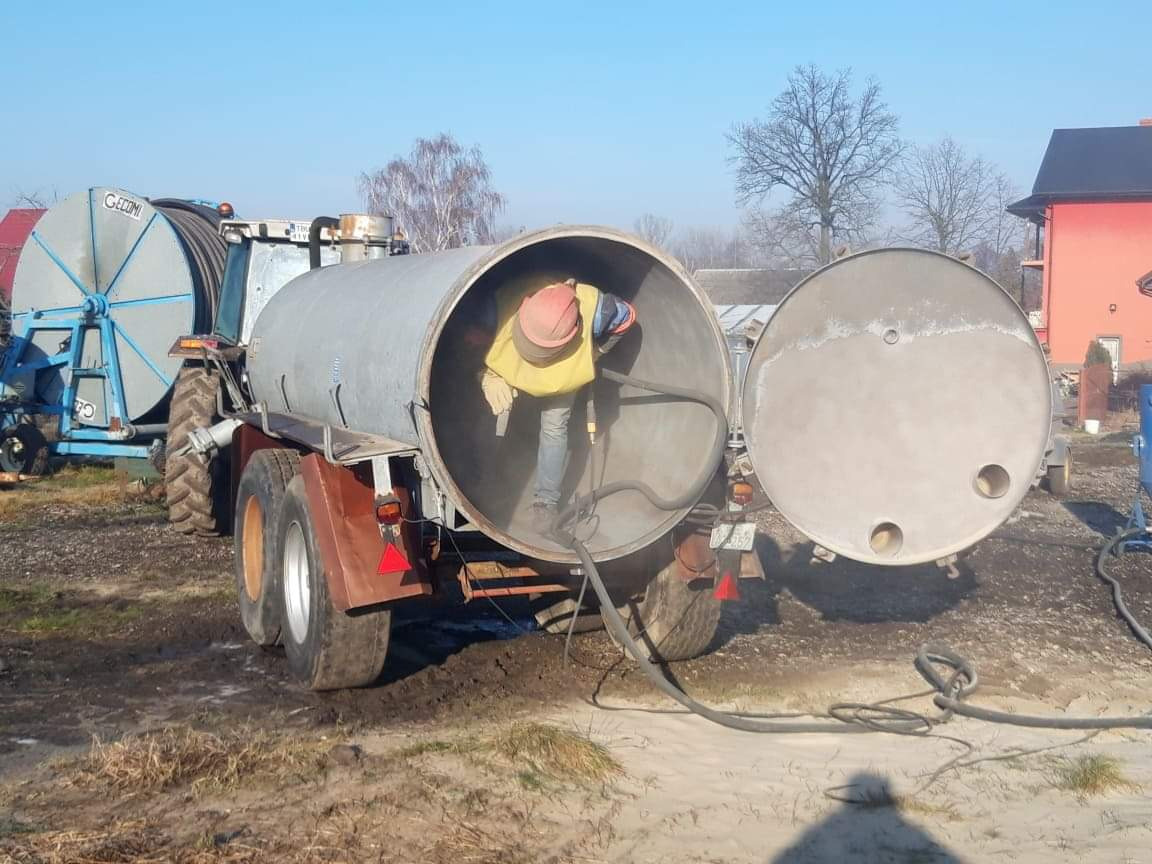 Pracownik w kombinezonie ochronnym wewnątrz otwartego zbiornika na przyczepie, czyszczący jego wnętrze za pomocą węża ciśnieniowego w plenerze.