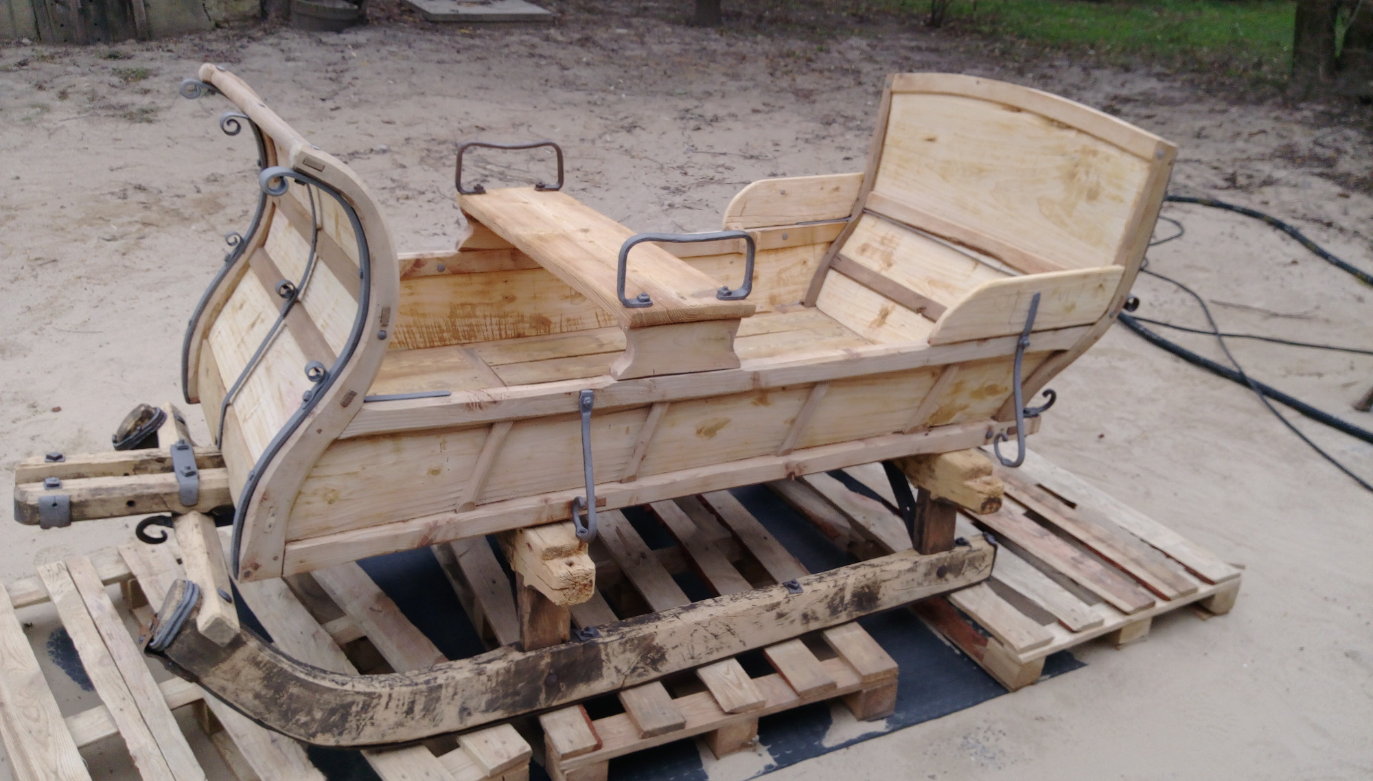 Surowa, drewniana konstrukcja sań na palecie, z metalowymi elementami łączącymi i uchwytami, w trakcie budowy lub renowacji.