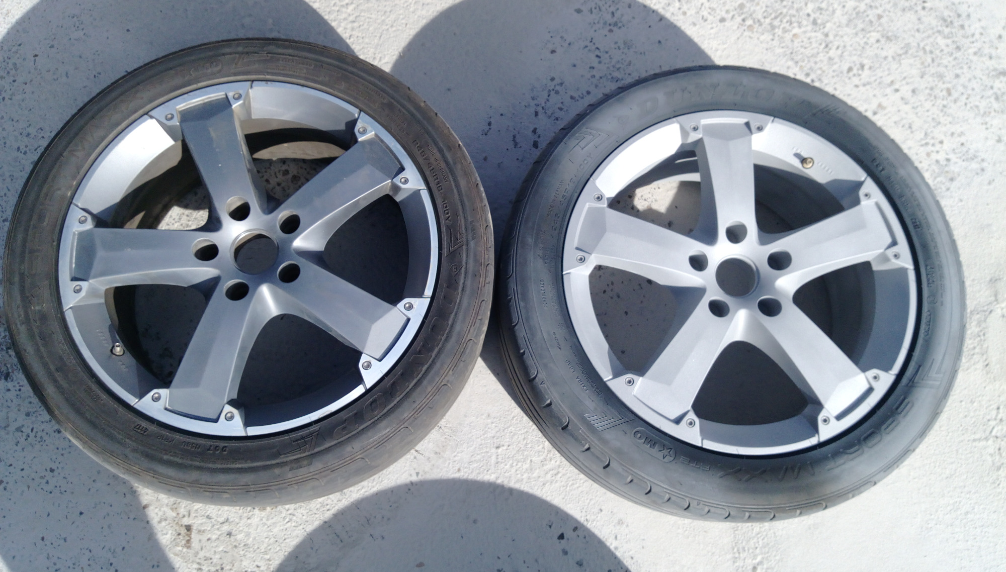 Dwa srebrne felgi aluminiowe z oponami leżące na betonowej powierzchni, widoczne ślady użytkowania.
