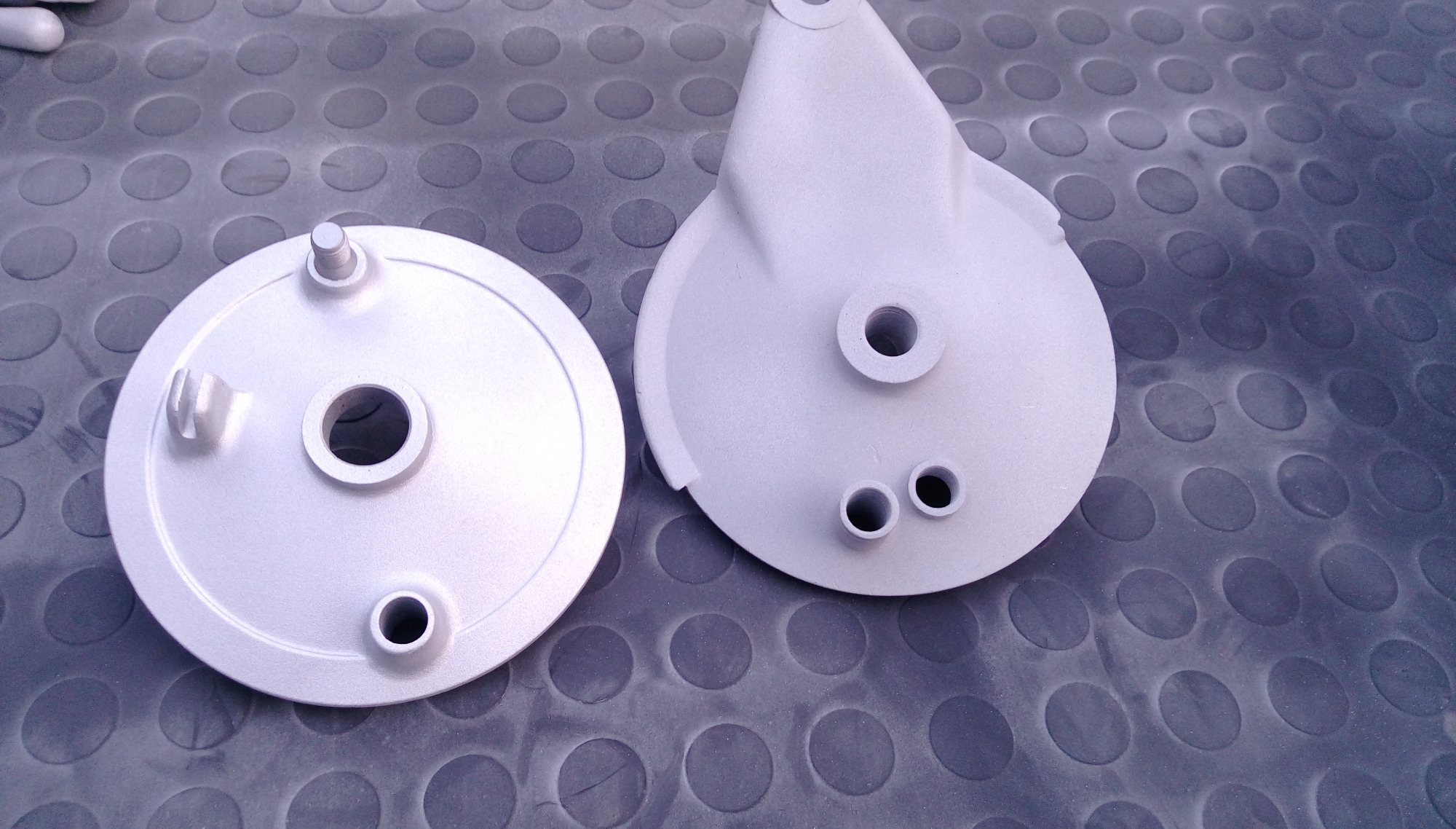 Dwie piasty hamulcowe motocyklowe, po obróbce, leżą na gumowej macie z wytłaczanymi kółkami. Piasty mają srebrzysto-szary matowy odcień.