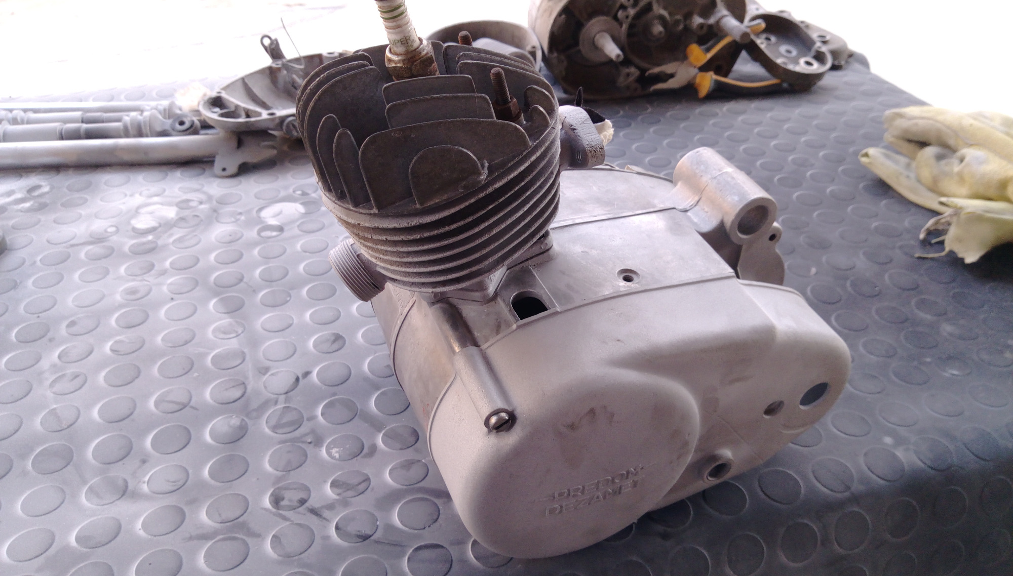Szary silnik motocyklowy po piaskowaniu, widoczne ślady po procesie, na tle gumowej maty warsztatowej, inne części silnika w tle.
