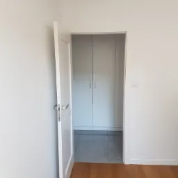 Biały anemostat sufitowy widoczny nad otwartymi drzwiami do garderoby z szarymi płytkami podłogowymi i drewnianą podłogą na pierwszym planie.