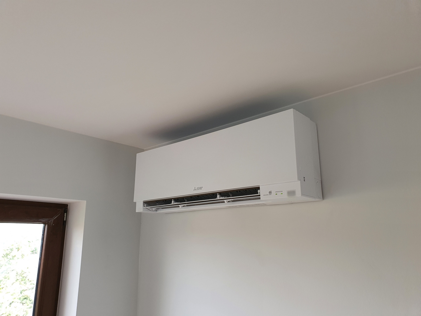 Biała jednostka klimatyzacji Mitsubishi zainstalowana na ścianie w jasnym pomieszczeniu w Grójcu, widoczna z boku okna.