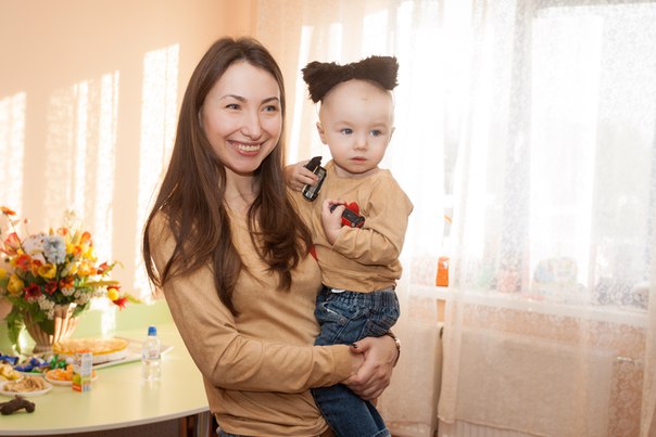 Uśmiechnięta kobieta trzyma na rękach małego chłopca w brązowej bluzce i jeansach, oboje ubrani w podobne stroje, w tle stół z kwiatami i przekąskami, jasne wnętrze z oknem.