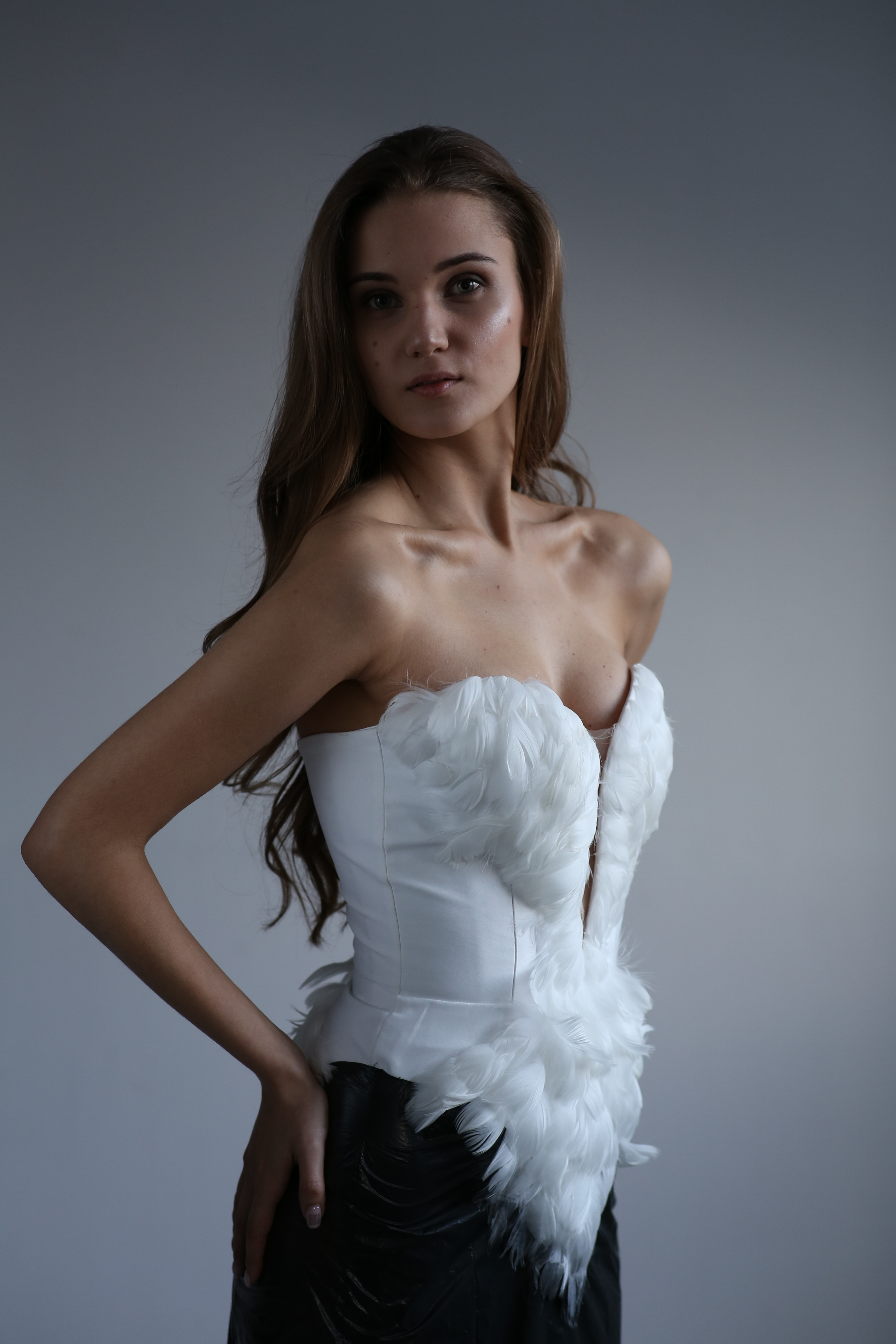 Modelka prezentuje unikatowy, biały gorset z piórami, wykonany w szwalni w Warszawie, z czarnym, skórzanym dołem, na jednolitym szarym tle.