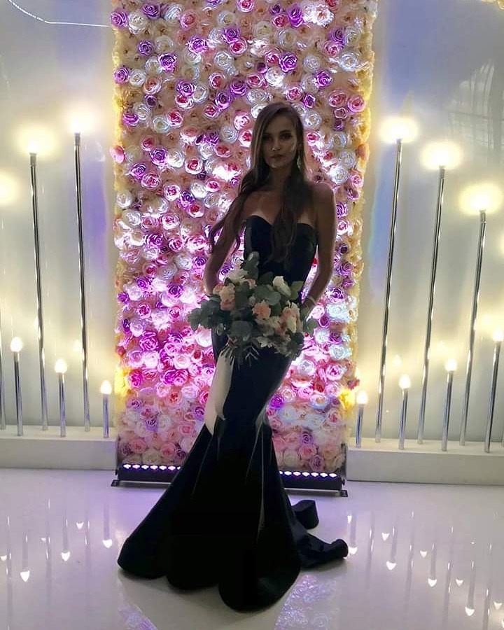 Elegancka kobieta w czarnej, dopasowanej sukni wieczorowej z bukietem kwiatów na tle ściany z róż i dekoracyjnych lamp.