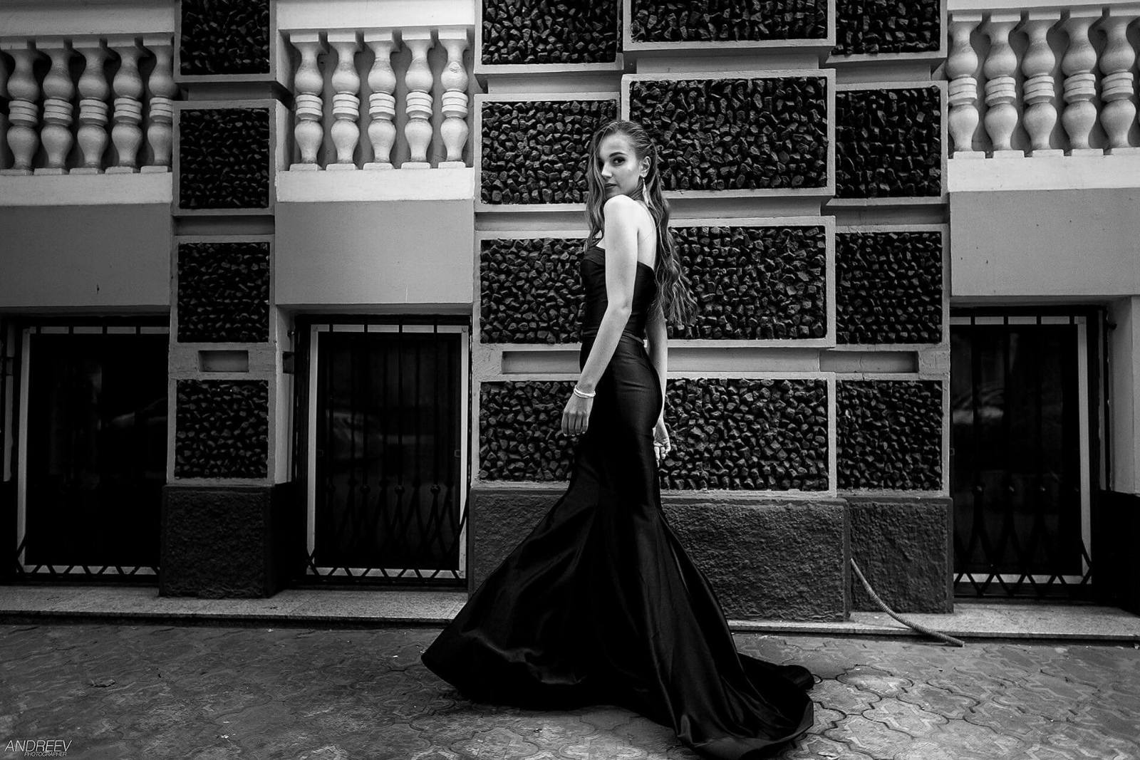 Czarno-białe zdjęcie kobiety w długiej, czarnej sukni wieczorowej, odwróconej przez ramię, na tle dekoracyjnej ściany z balustradami i kamiennymi panelami.
