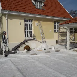 Malowanie elewacji domu jednorodzinnego w trakcie realizacji, widoczny pracownik w kombinezonie, zabezpieczone okna taśmą, rozłożona folia ochronna, drabina oparta o ścianę.