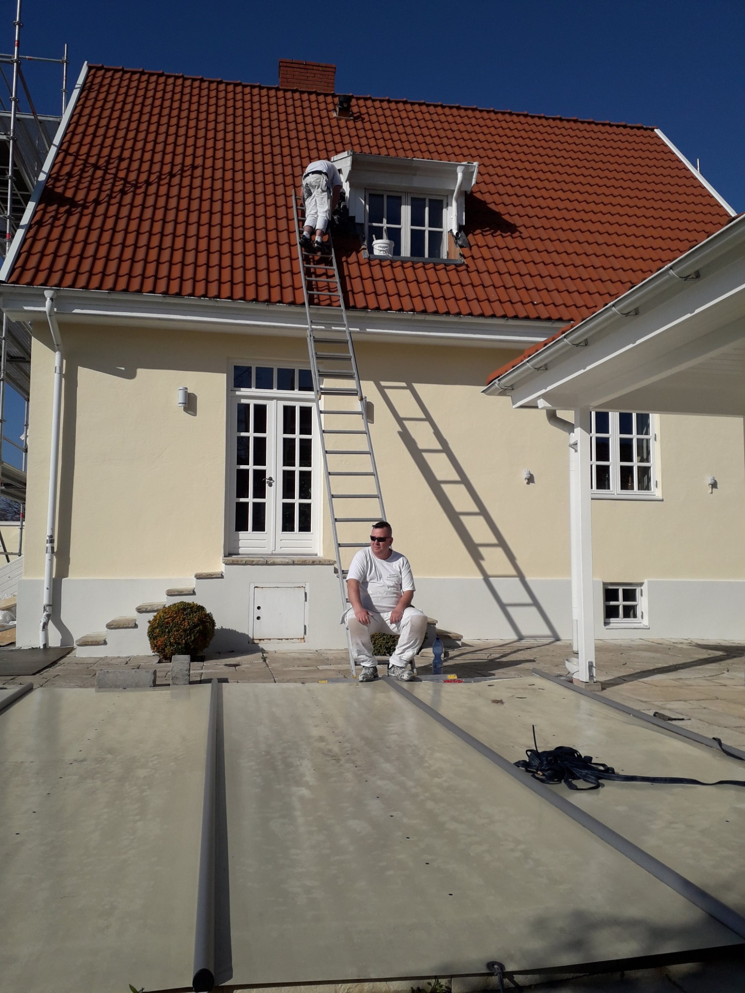 Dwóch mężczyzn w białych kombinezonach podczas odnawiania elewacji budynku z czerwoną dachówką; jeden maluje pod okapem na drabinie, drugi siedzi przed budynkiem.