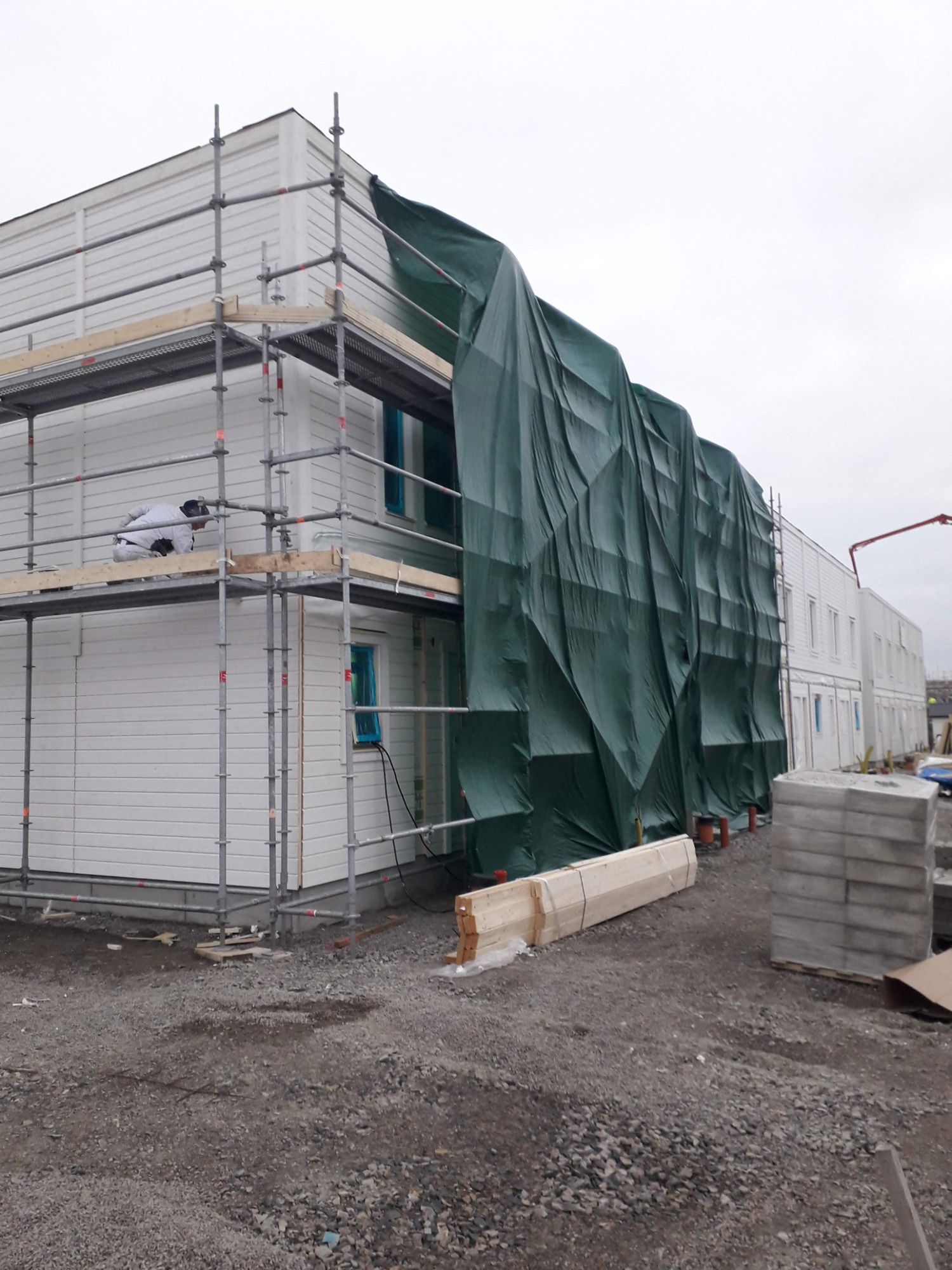 Budynek w trakcie prac wykończeniowych, zabezpieczony zieloną plandeką, widoczne rusztowania i pracownik w kombinezonie ochronnym na wysokości.