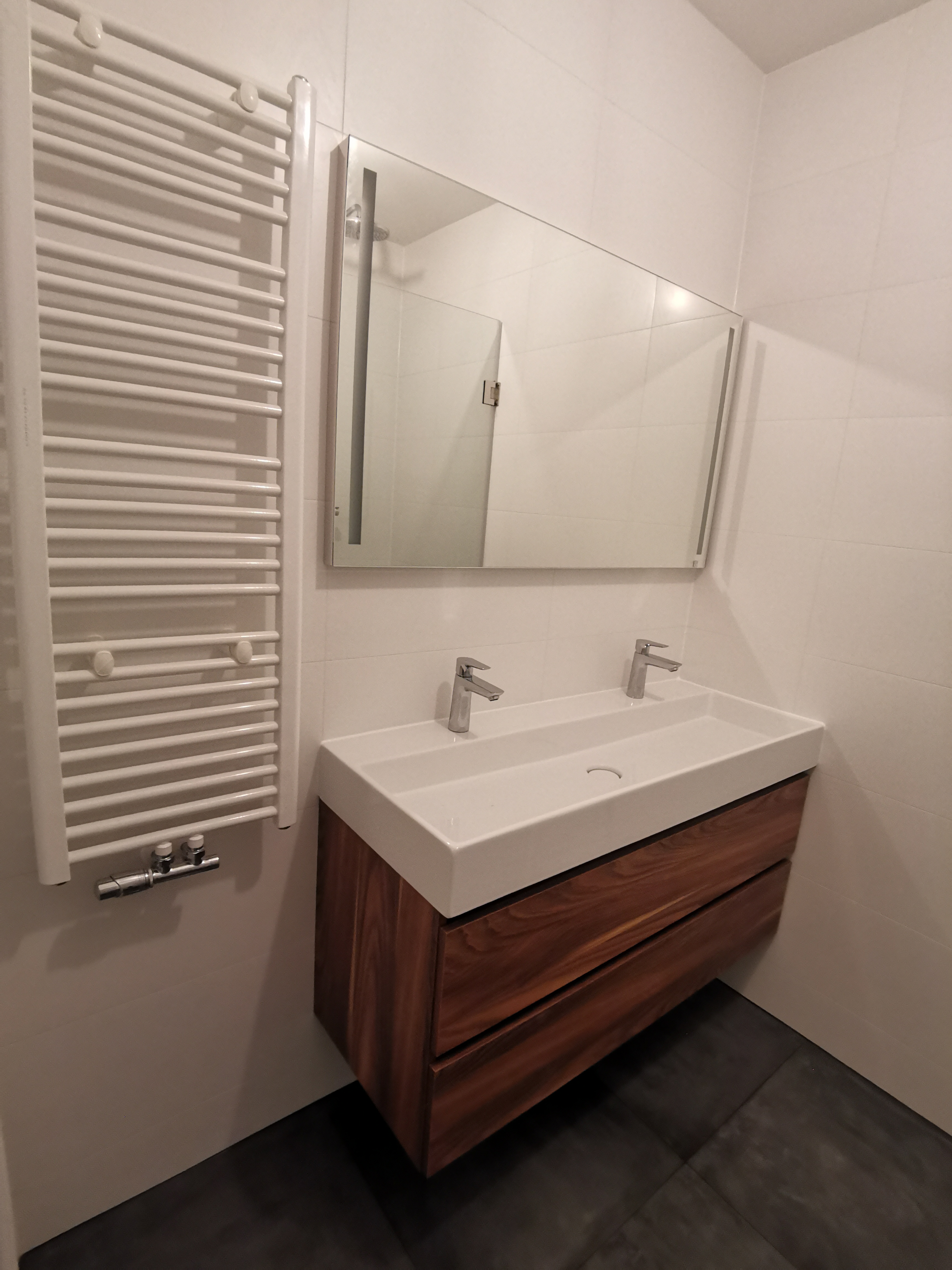 Nowoczesna łazienka z podwójną umywalką nablatową na drewnianej szafce, podświetlane lustro i biały grzejnik drabinkowy na tle białych płytek.