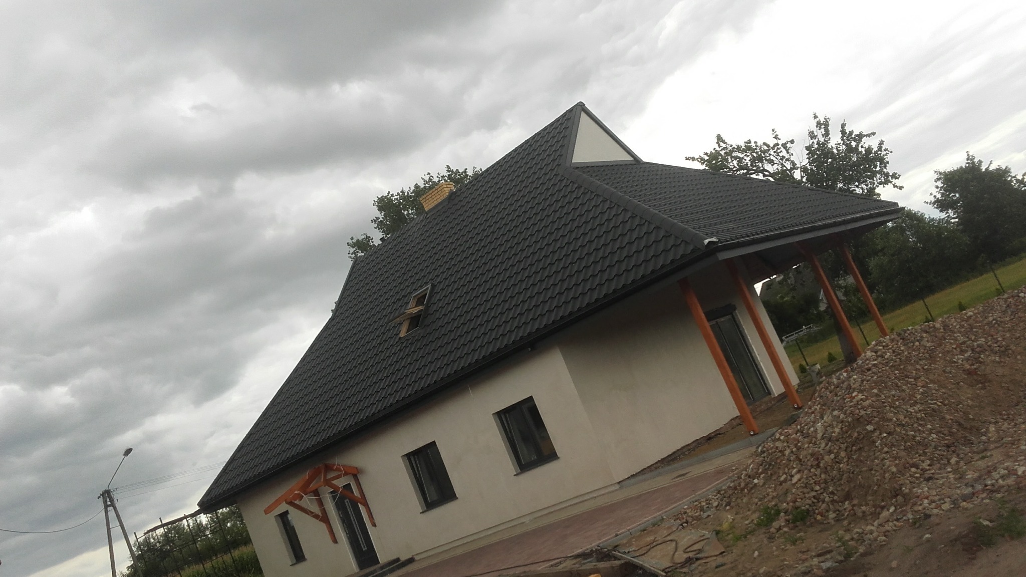 Dom jednorodzinny z ciemną dachówką modułową, widoczne okno dachowe, świeżo otynkowana elewacja i sterta kamieni na posesji, pochmurne niebo w tle.