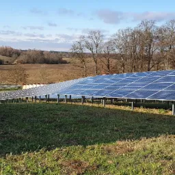 Rozległa instalacja paneli słonecznych na łące z widokiem na pola uprawne i drzewa w tle, w słoneczny dzień.