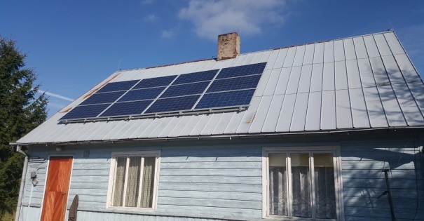 Panele słoneczne na blaszanym dachu drewnianego domu z kominem, widoczne z poziomu gruntu w słoneczny dzień.