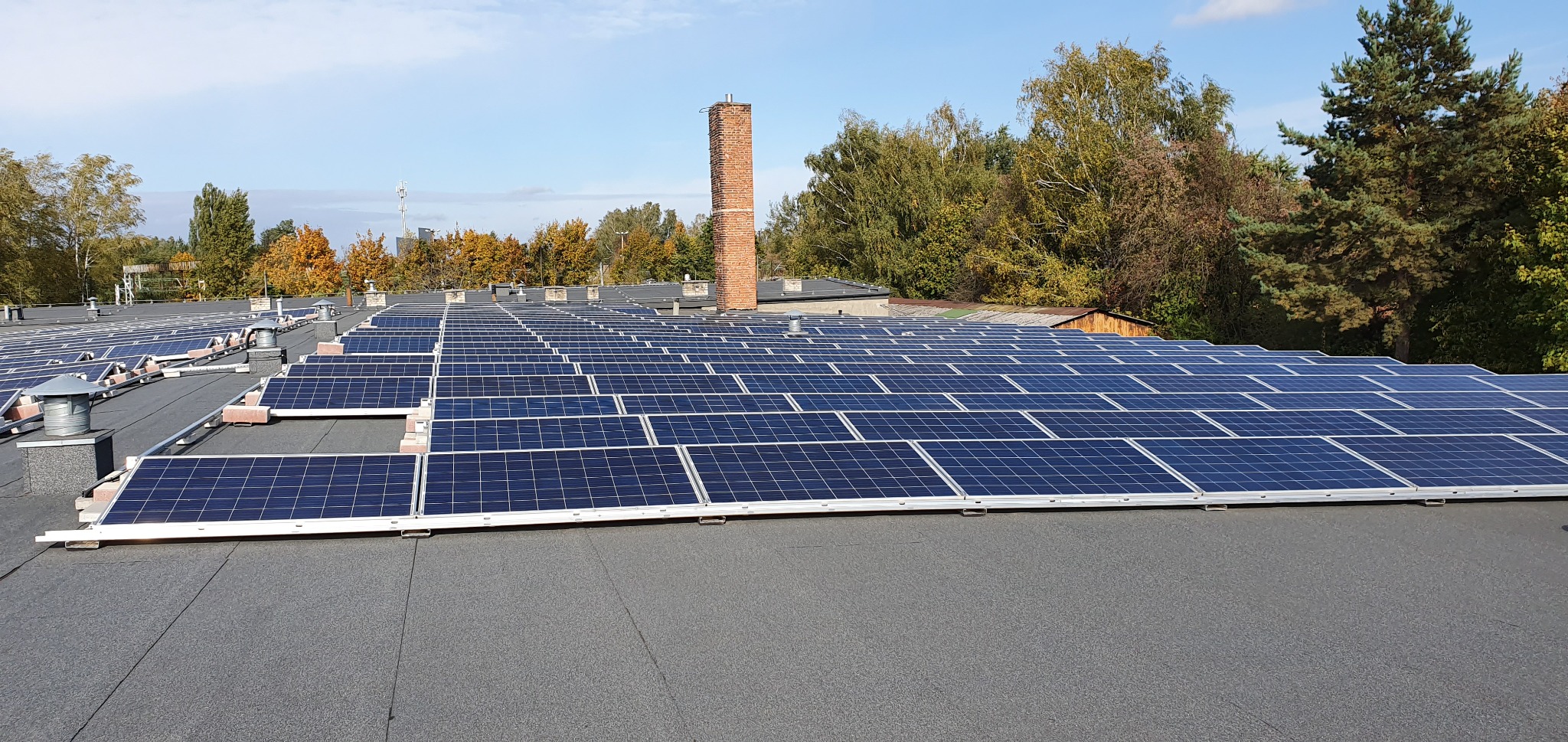 Rozległa instalacja paneli słonecznych na płaskim dachu budynku z kominem w tle, widok z góry.