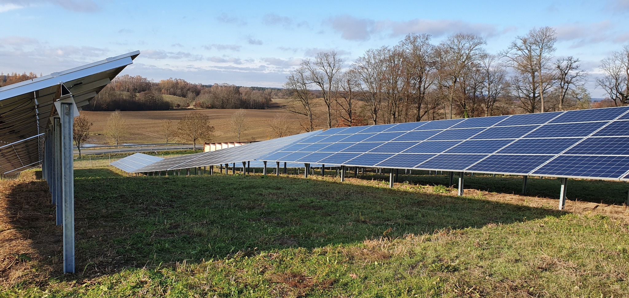 Rozległa instalacja paneli słonecznych na łące z widokiem na pola uprawne i drzewa w tle, w słoneczny dzień.