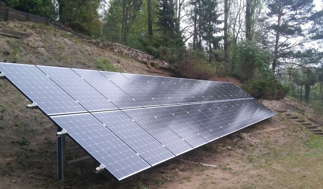 Instalacja fotowoltaiczna na zboczu wzgórza, panele ustawione pod kątem, widoczne metalowe wsporniki, w tle drzewa i kamienne schody.