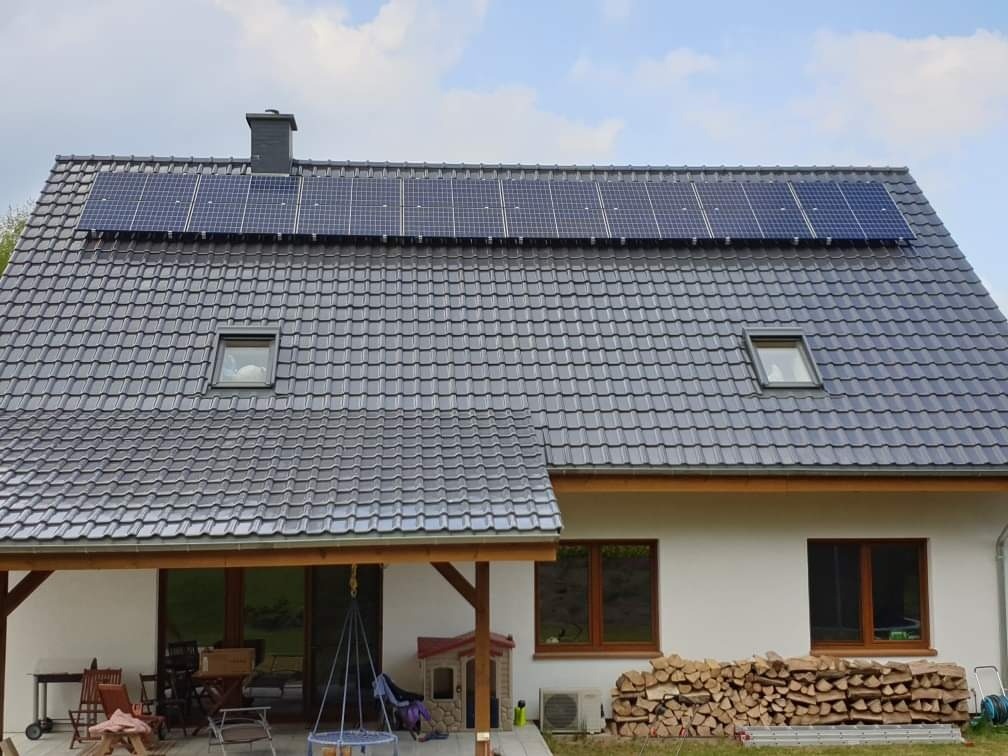 Dom jednorodzinny z szarą dachówką i panelami słonecznymi na dachu, widoczne okna dachowe, drewniana wiata i ułożone drewno kominkowe.