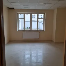 Pusty pokój biurowy po remoncie, świeżo pomalowane ściany w odcieniach beżu, gładka wylewka na podłodze, okno z widokiem na budynek, biały grzejnik, sufit kasetonowy z brakującym panelem.