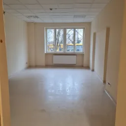 Puste, jasne pomieszczenie biurowe po remoncie, z widokiem na żółty tramwaj za oknem i śladami po pracach wykończeniowych na podłodze.