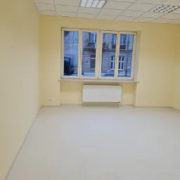 Pusty pokój biurowy z kremowymi ścianami, jasną podłogą i oknem z widokiem na budynek mieszkalny, po remoncie.