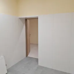 Wnętrze pomieszczenia z wyłożonymi białymi płytkami ceramicznymi na ścianach do połowy wysokości i szarymi płytkami na podłodze, widoczne drzwi do innego pomieszczenia i grzejnik.