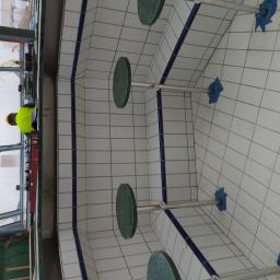 Pol-Ros - Wnętrze basenu w trakcie budowy z białymi płytkami, niebieskimi akcentami i zamontowanymi okrągłymi siedziskami, pracownik w żółtej kamizelce poziomuje elementy konstrukcyjne.