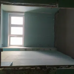 Wnętrze w trakcie remontu: ściany wyłożone płytami kartonowo-gipsowymi, widoczne okno i surowy beton na podłodze, ujęcie z perspektywy wejścia.