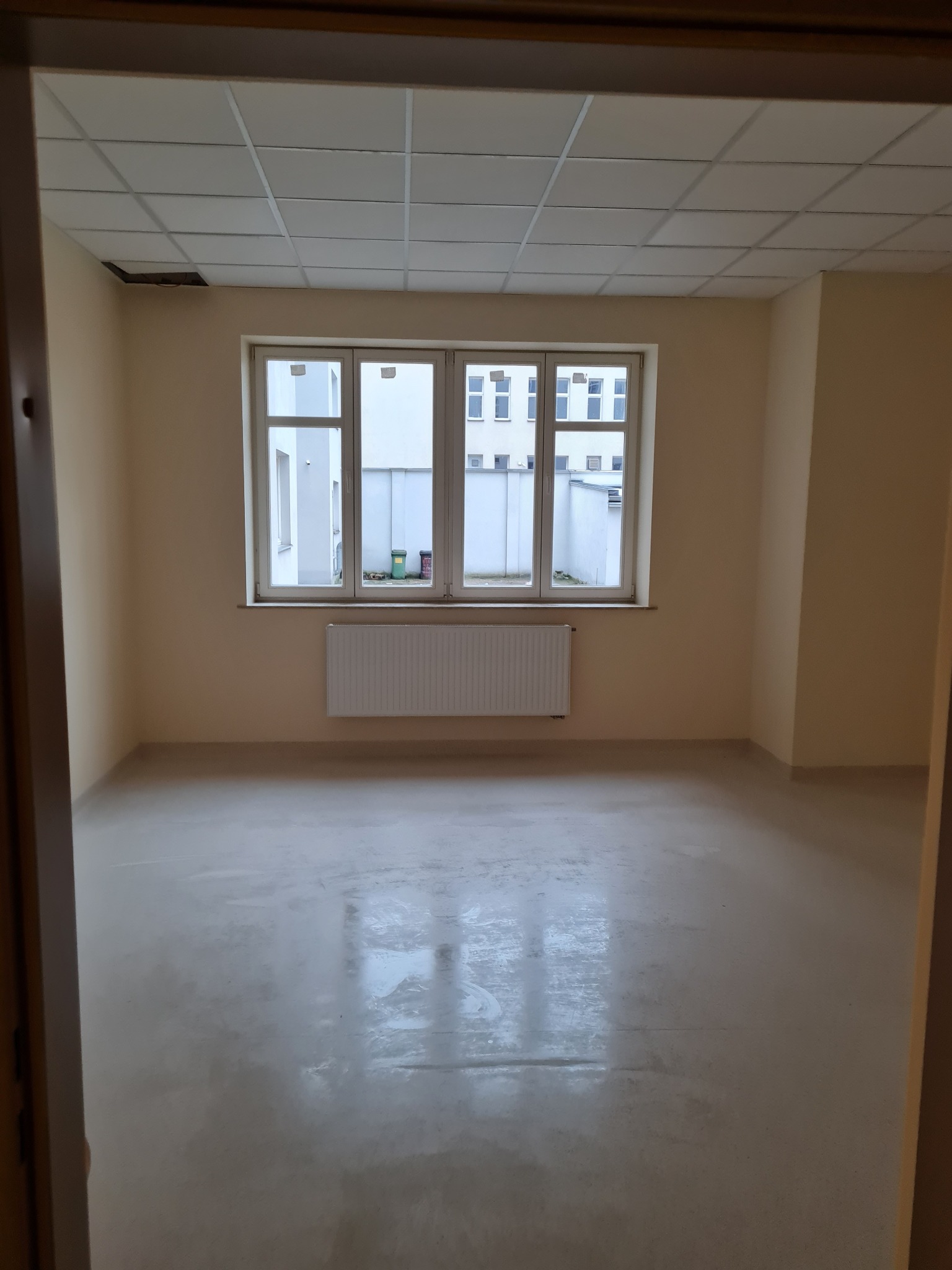 Pusty pokój biurowy po remoncie, świeżo pomalowane ściany w odcieniach beżu, gładka wylewka na podłodze, okno z widokiem na budynek, biały grzejnik, sufit kasetonowy z brakującym panelem.
