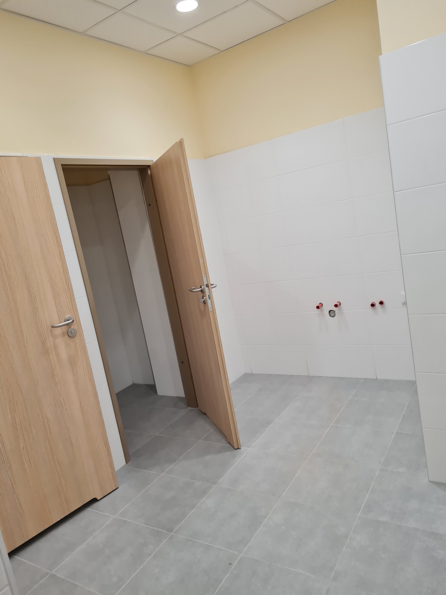 Wnętrze publicznej toalety z kabinami, wykończone białymi płytkami i szarą podłogą, widoczne wystające rury instalacji wodnej.