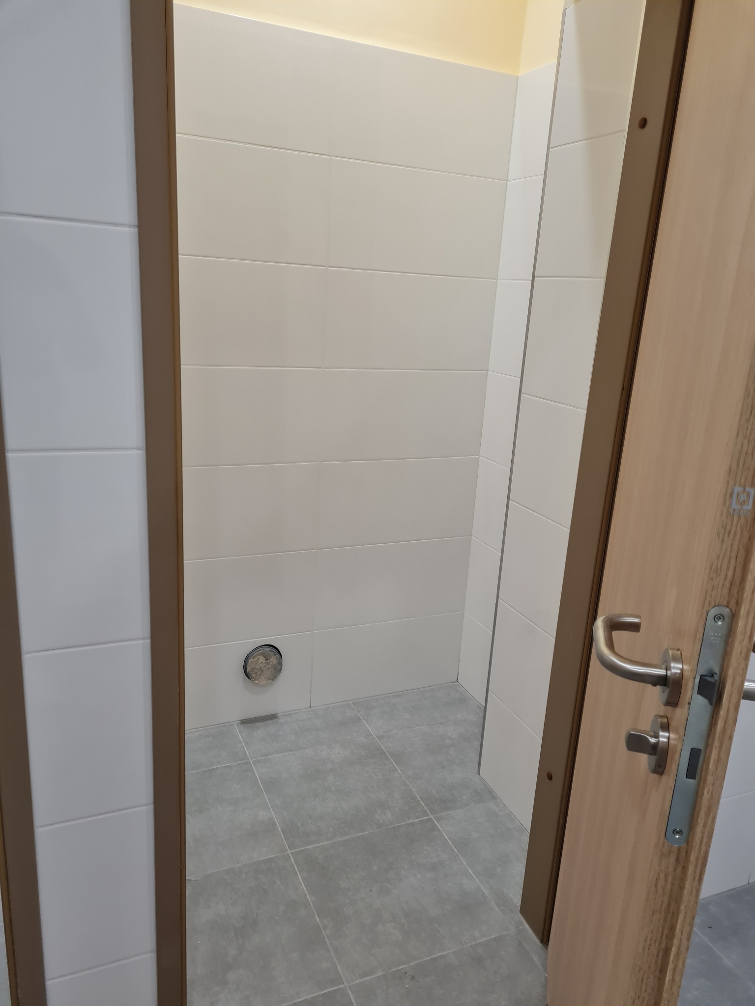 Wykończona łazienka z jasnymi płytkami na ścianach i szarą podłogą, widok przez otwarte drzwi z drewnianą ościeżnicą i srebrną klamką. Otwór wentylacyjny w ścianie.