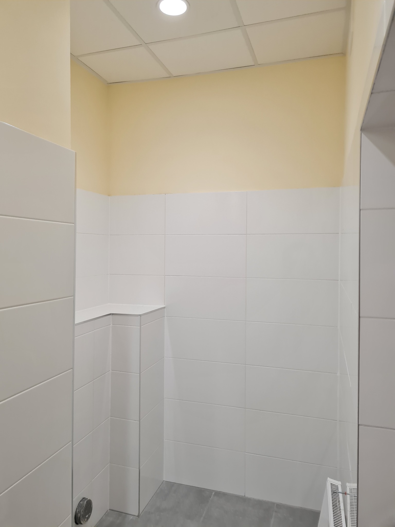 Wnętrze łazienki po remoncie: białe płytki na ścianach do połowy wysokości, kremowa farba powyżej, szara podłoga, widoczny fragment grzejnika i odpływ.