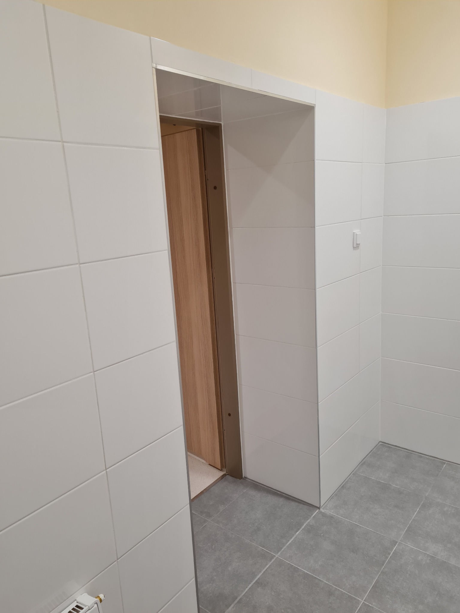 Wykończona łazienka z białymi płytkami na ścianach i szarymi na podłodze, widoczne ościeżnice drewniane drzwi, pomalowane na żółto górne partie ścian.