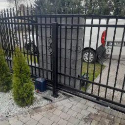 Czarna, metalowa brama wjazdowa przesuwna z automatyką, widoczne dwa małe drzewka iglaste i częściowo biały samochód dostawczy w tle.