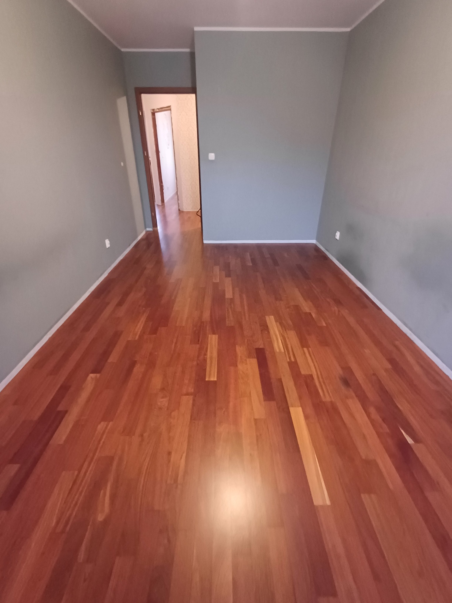 Wypolerowany parkiet w odcieniach czerwieni, widok z niskiej perspektywy, szare ściany i otwarte drzwi w tle.