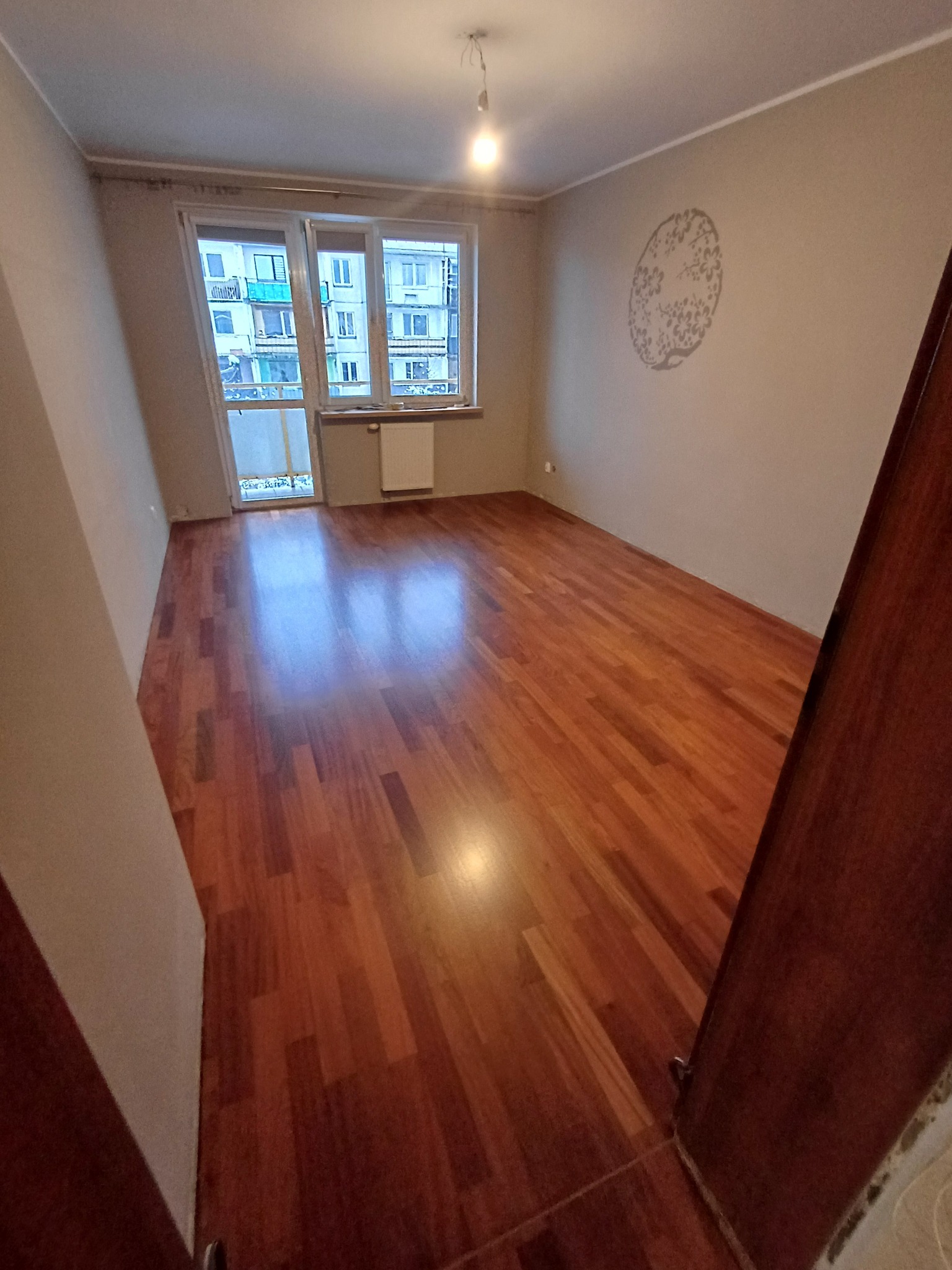 Wypolerowany parkiet w pustym pokoju z beżowymi ścianami, widok zza otwartych drzwi, na ścianie dekoracyjny wzór.