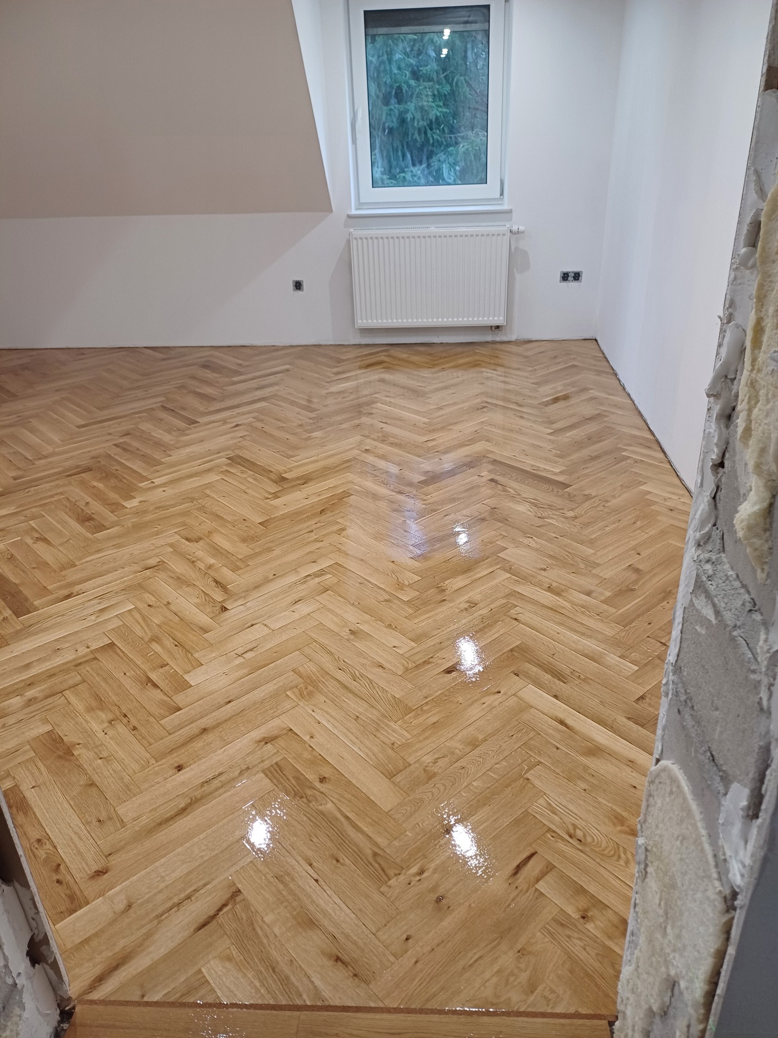 Świeżo polakierowany parkiet ułożony w jodełkę w jasnym pomieszczeniu z oknem, widoczne ślady po remoncie na ścianie po prawej stronie.