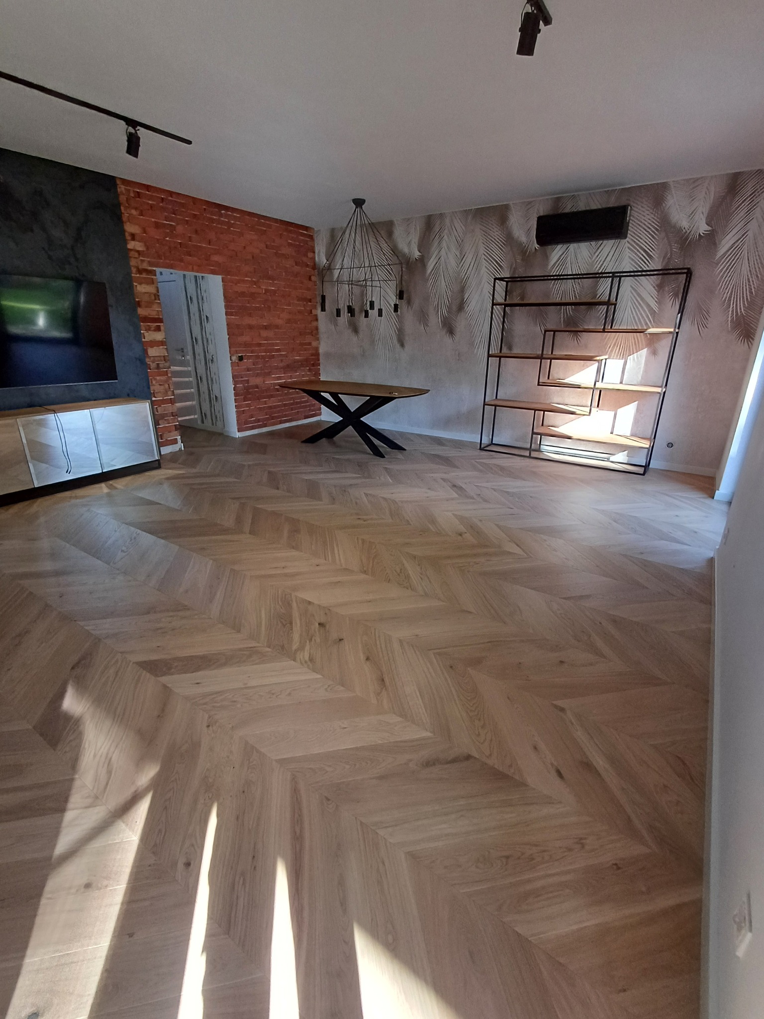 Jasna, olejowana podłoga z parkietu ułożonego we wzór jodły francuskiej, widok na salon z ceglaną ścianą, nowoczesnym regałem i lampą.