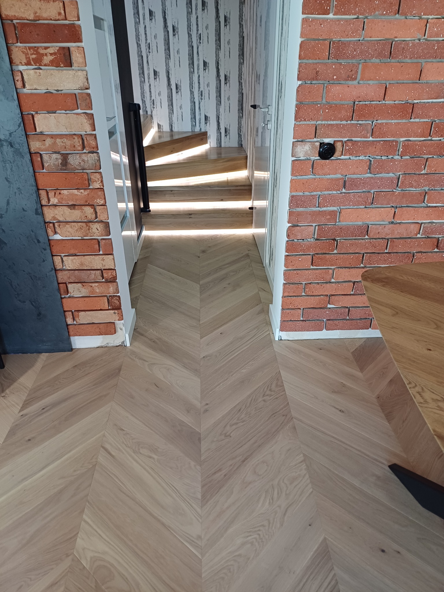 Olejowana podłoga z parkietu ułożonego we wzór jodły francuskiej, widok z góry w kierunku schodów z drewnianymi stopniami, ściana z czerwonej cegły po prawej stronie.