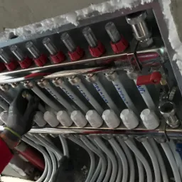 Rozdzielacz ogrzewania podłogowego w metalowej obudowie, z podłączonymi rurami oznaczonymi nazwami pomieszczeń, instalowany przez osobę w rękawicy roboczej.