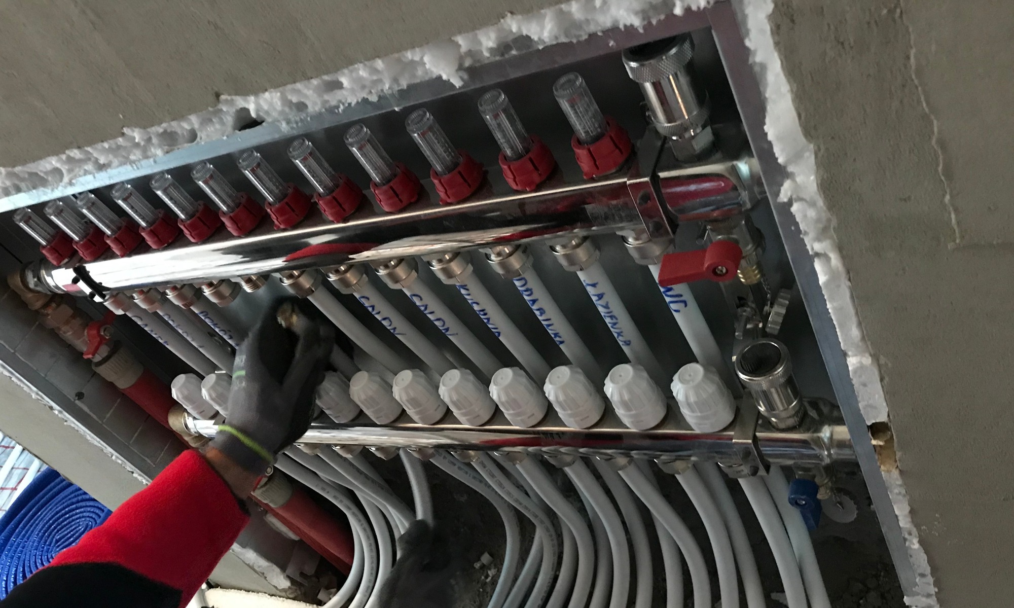 Rozdzielacz ogrzewania podłogowego w metalowej obudowie, z podłączonymi rurami oznaczonymi nazwami pomieszczeń, instalowany przez osobę w rękawicy roboczej.