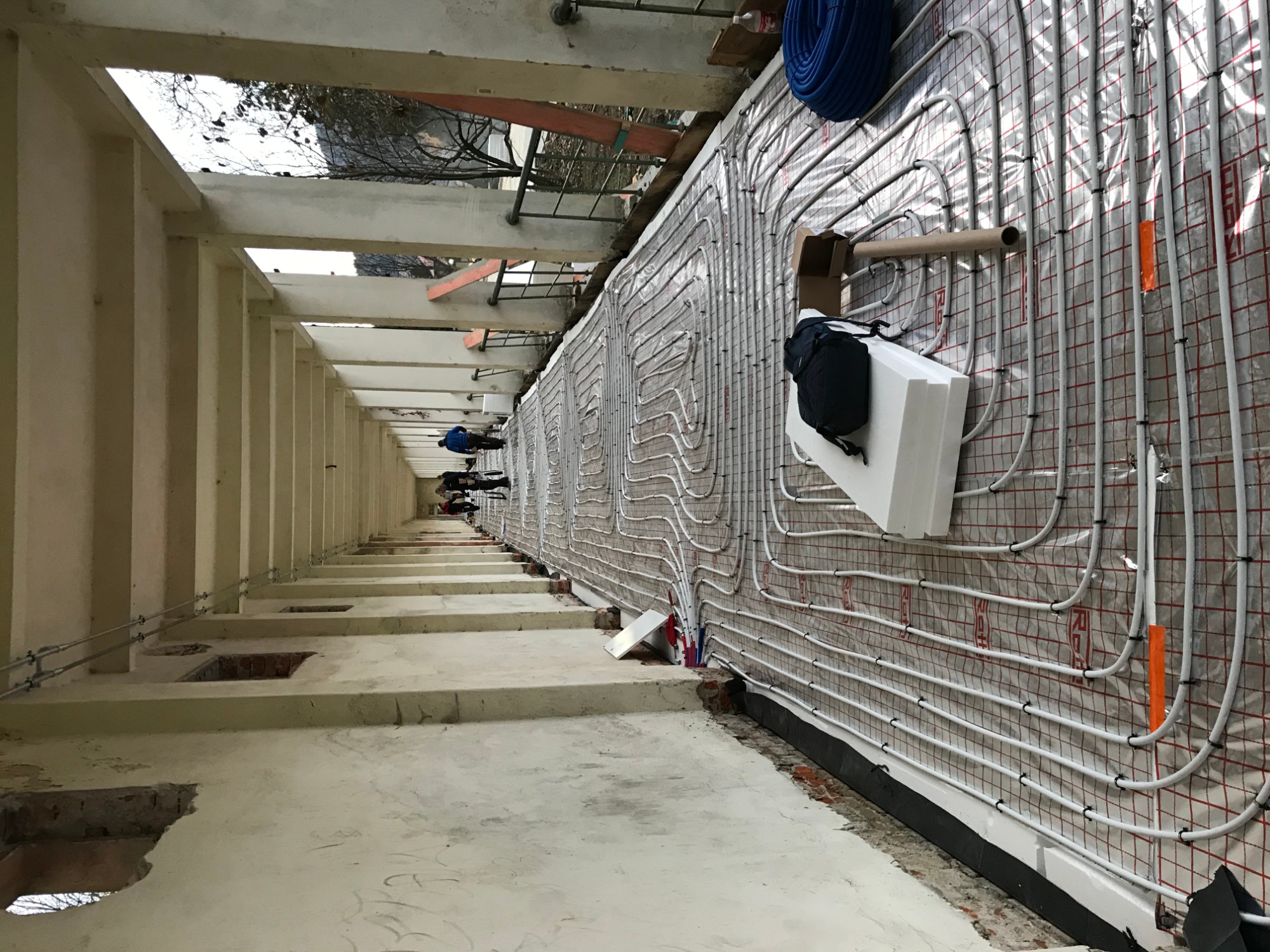 Montaż ogrzewania podłogowego: białe rurki ułożone równolegle na folii z czerwoną kratką, widoczne elementy izolacji, czarny plecak i niebieski zwój rur, w tle surowy betonowy strop budynku...