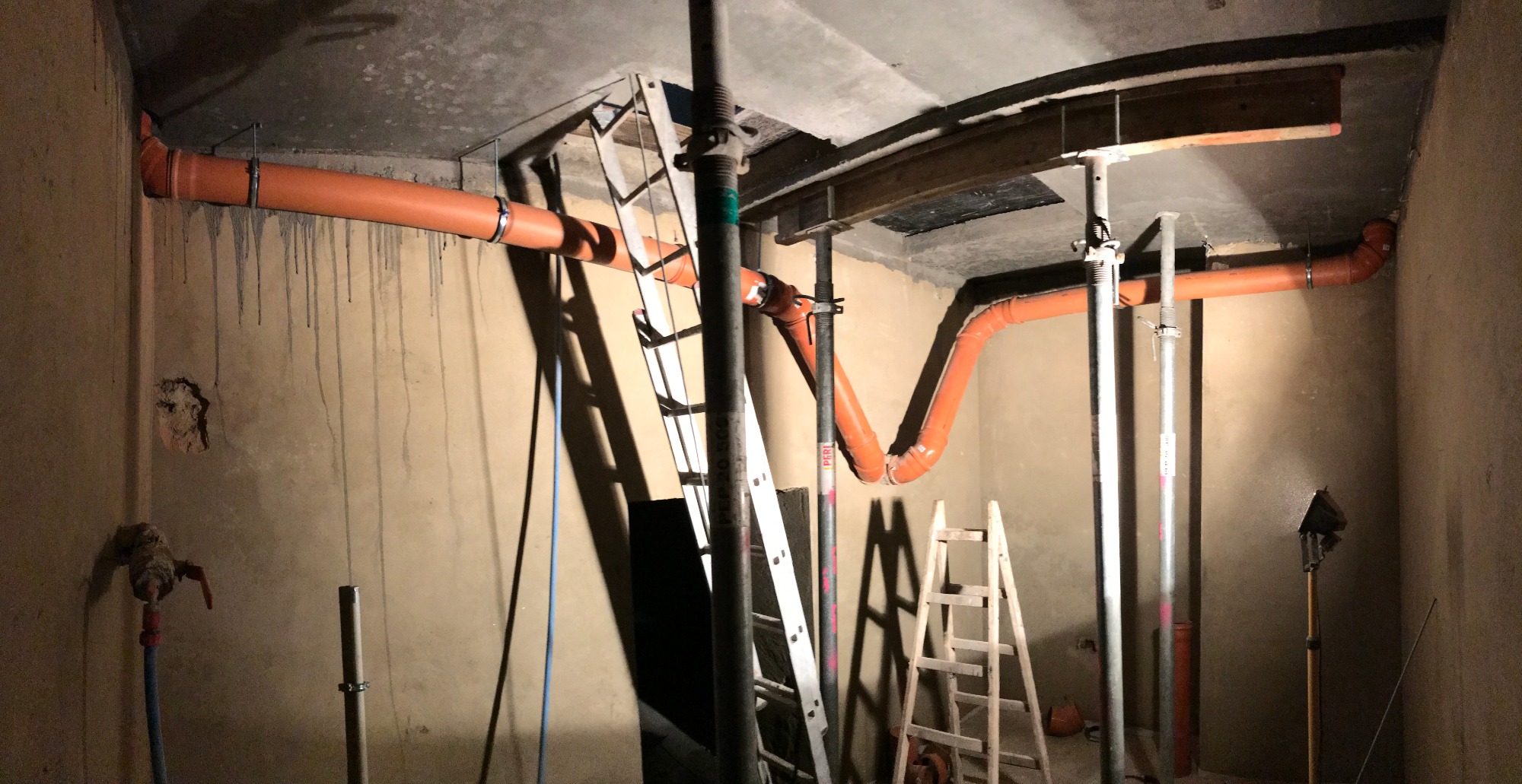 Pomarańczowe rury kanalizacyjne zamontowane pod sufitem w surowym wnętrzu, wsparte na metalowych podporach i drabinkach, ściany z zaciekami.