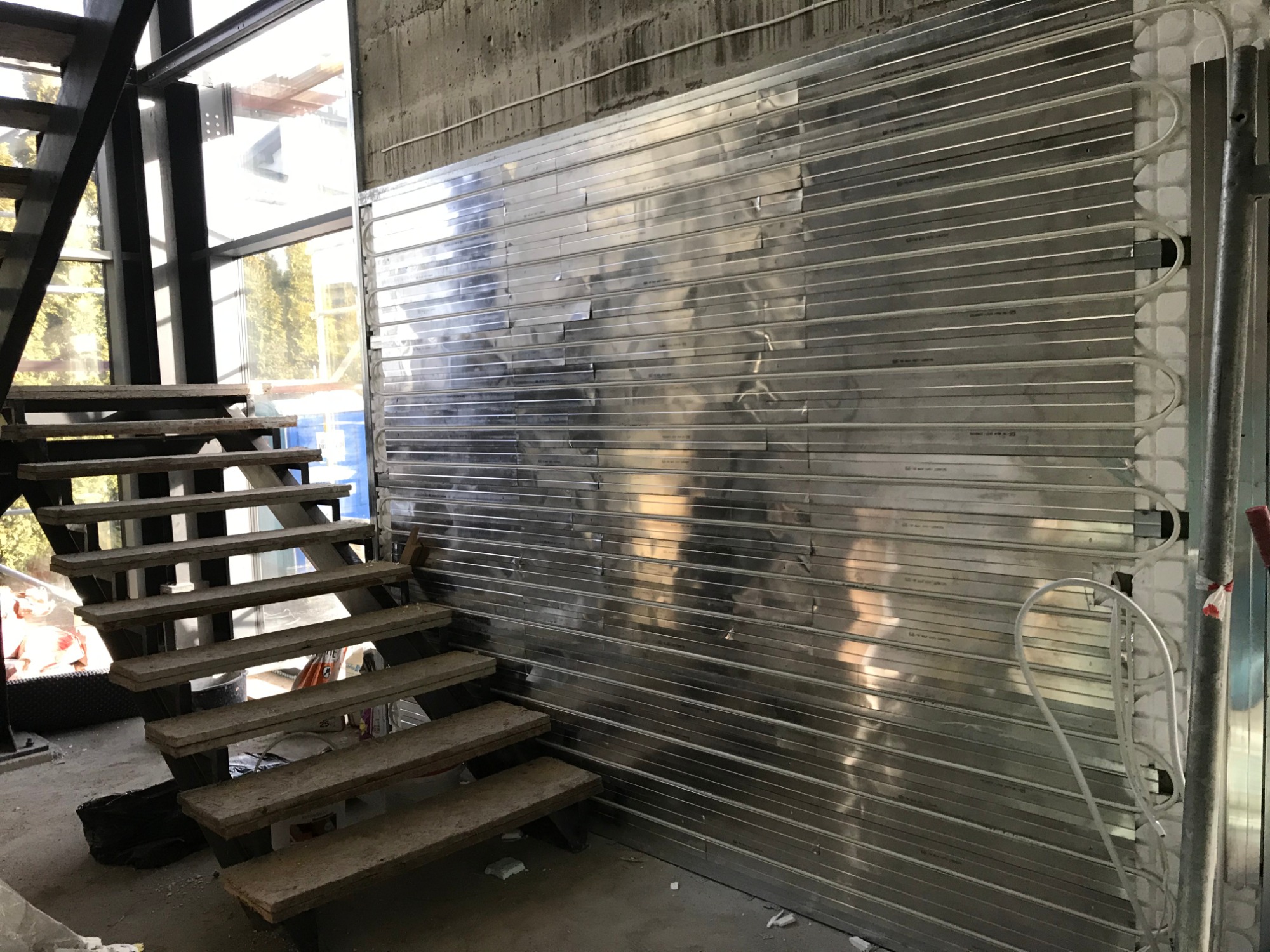 Ściana z panelami grzewczymi w surowym stanie, z widocznymi rurami doprowadzającymi, obok schody stalowe i betonowa ściana w tle