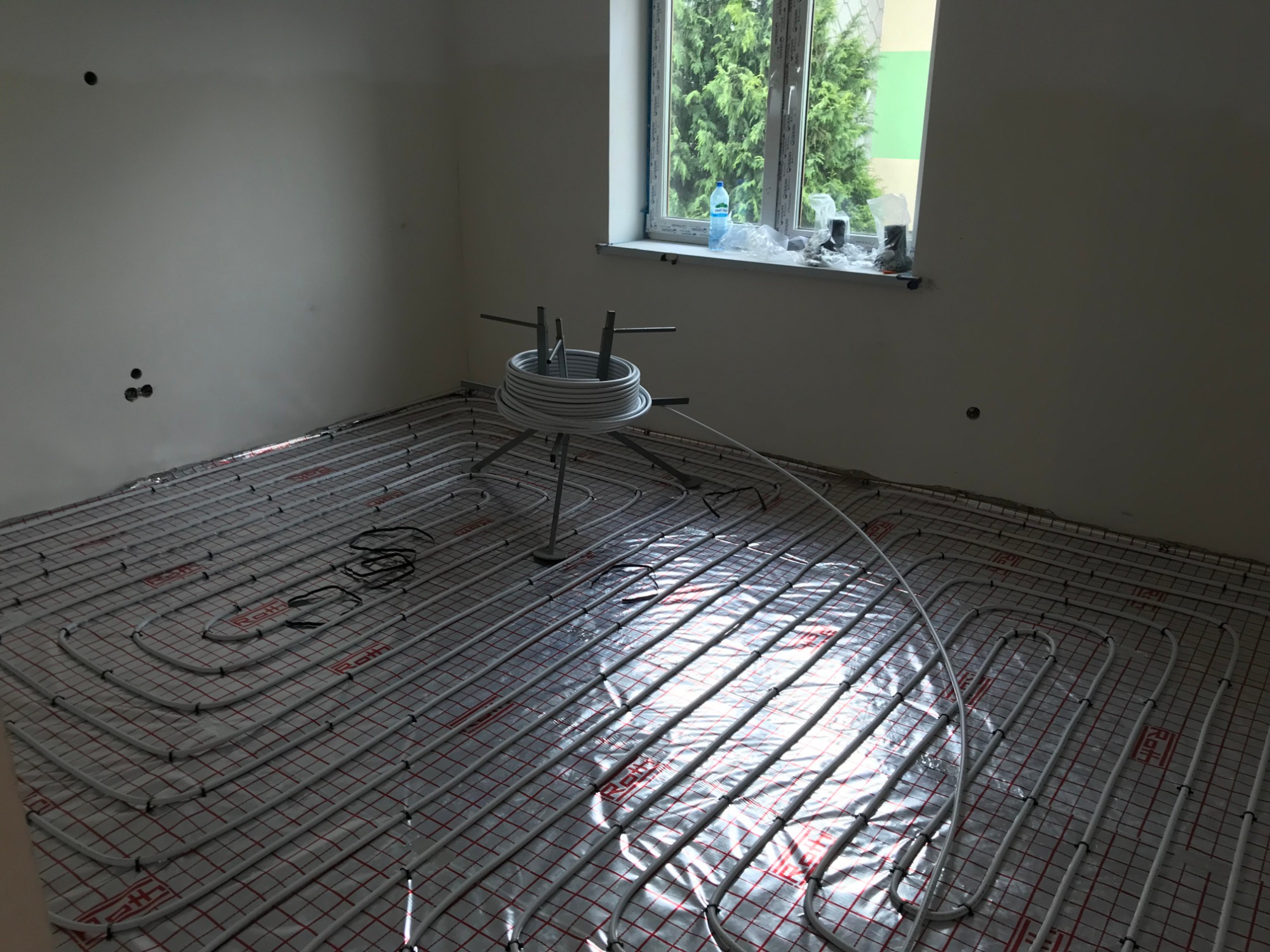 Rozłożona instalacja ogrzewania podłogowego na folii z siatką w pustym pomieszczeniu, widoczny zwój rur na metalowym stojaku, okno z widokiem na zieleń.