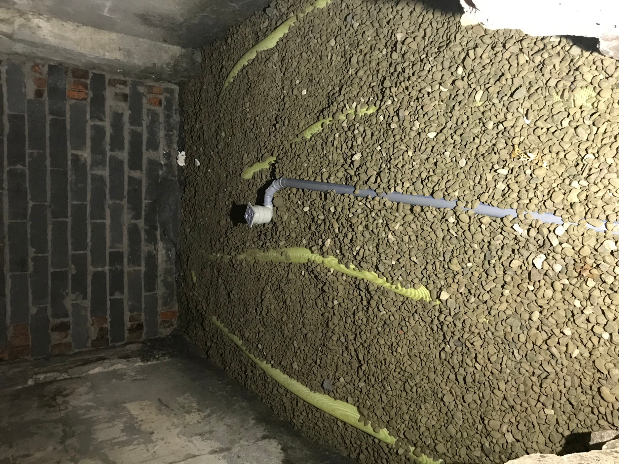 Ściana piwnicy pokryta warstwą izolacyjną z drenażem, widoczna rura odprowadzająca wodę i fragment ściany z cegły