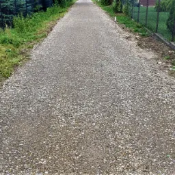 Długa, świeżo wysypana żwirowa ścieżka ogrodowa, wyznaczona sznurkiem, z widocznymi śladami po pracy, obrzeża porośnięte trawą i krzewami, w tle siatka ogrodzeniowa.